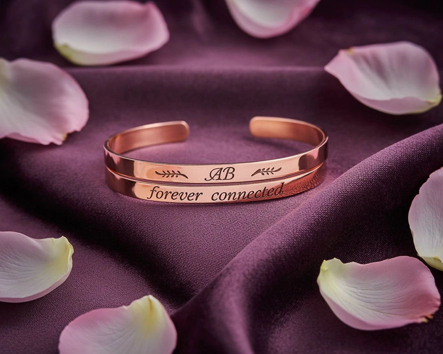 Matching engraved copper cuff bracelets on velvet fabric, featuring initials and meaningful messages, surrounded by rose petals.