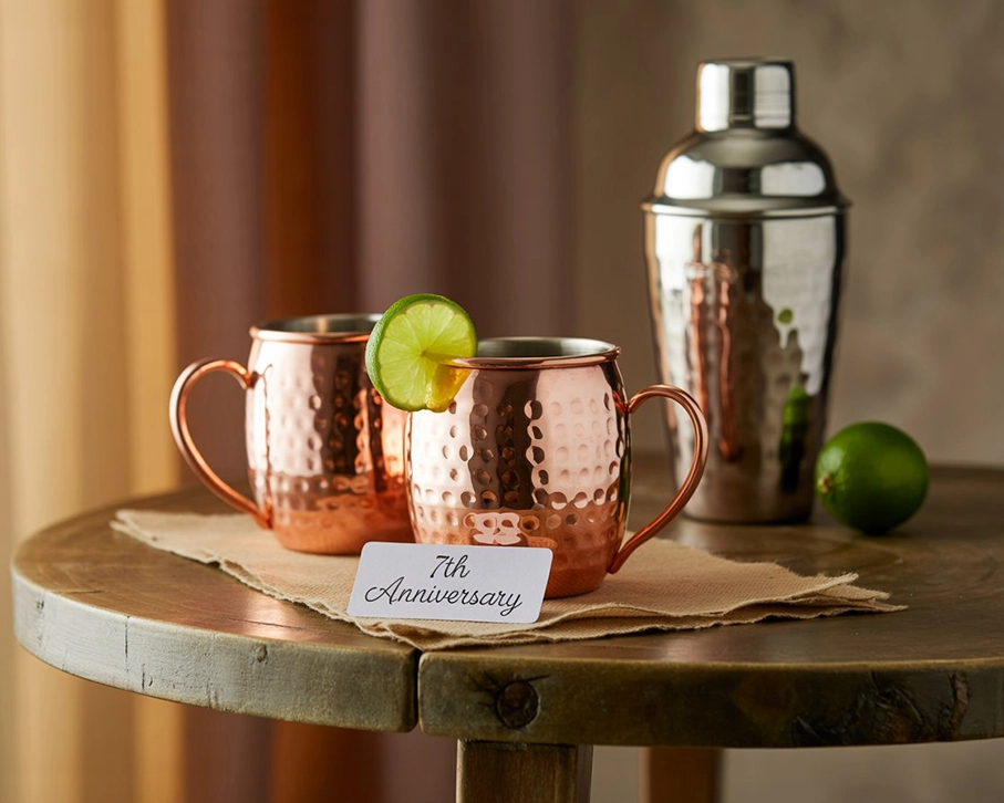 Hammered copper mugs with a cocktail shaker and lime garnish on a rustic table, styled as a 7th anniversary gift set.